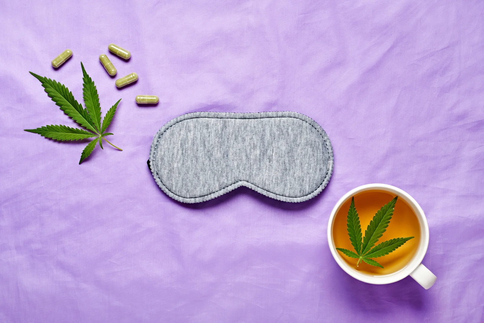 Flat lay with hemp leaves, capsules, tea cup, and sleep mask on lavender fabric, symbolizing natural remedies and cannabis for sleep support.
